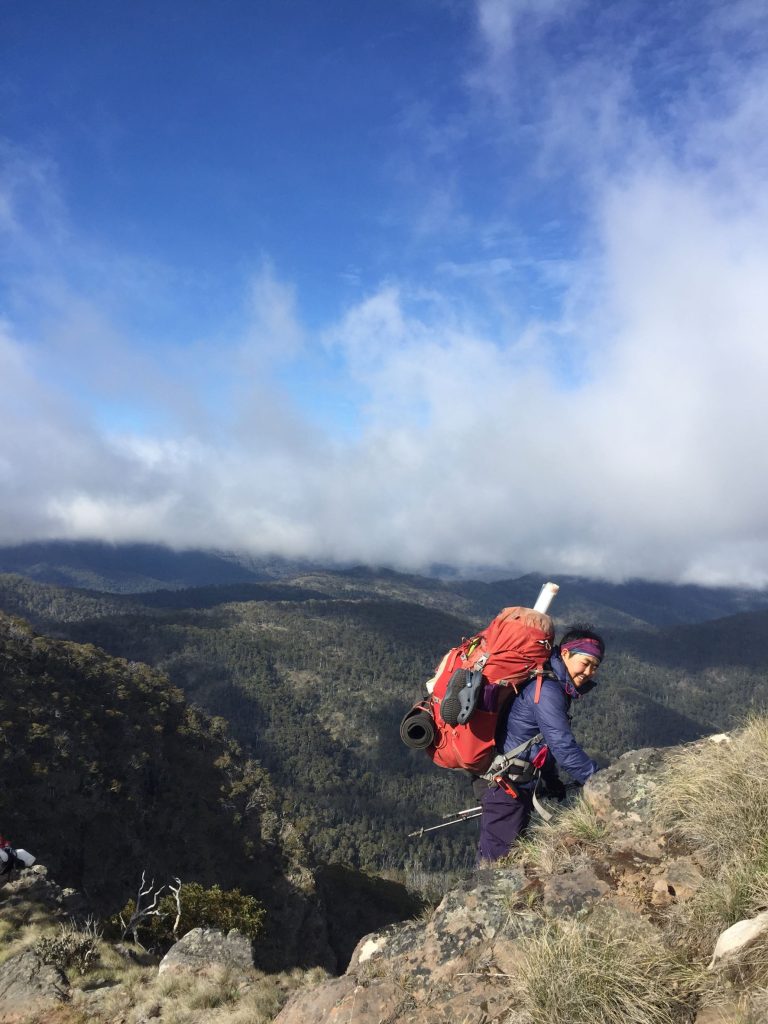 A woman with Chinese appearance with short black hair, wearing a headband, blue jacket and purple pants, carrying a red backpack with maps and equipment, climbing down a rocky outcrop, with a background of mountains and rolling clouds and blue sky.